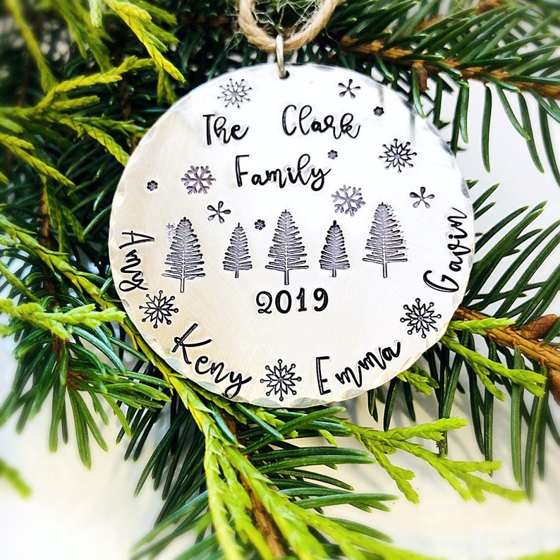 May include: Silver Christmas ornament with the text "The Clark Family 2019" and the names "Amy, Kenny, Emma, Gavin" around the edge. There are five stylized Christmas trees and snowflakes around the ornament.