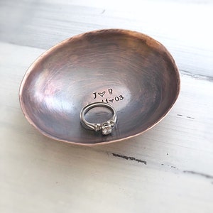 May include: A small, oval-shaped, hammered copper trinket dish with a dark, weathered patina. The interior is stamped with the text J heart B and 11 heart 03. A silver-toned ring with a central stone sits inside the bowl of the dish.