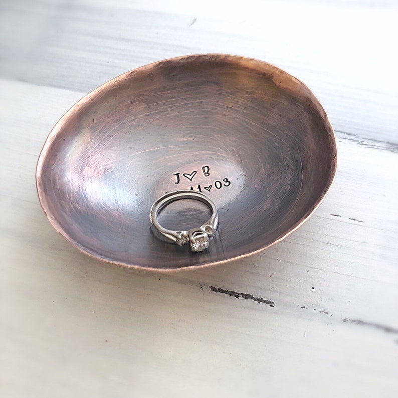 May include: A small, oval-shaped, hammered copper trinket dish with a dark, weathered patina. The interior is stamped with the text J heart B and 11 heart 03. A silver-toned ring with a central stone sits inside the bowl of the dish.