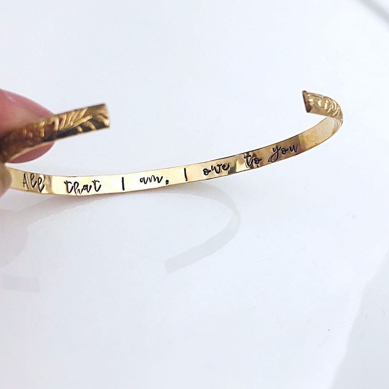 A delicate and minimalist 14 gold filled cuff bracelet featuring a floral pattern on the outside, with the words “All that I am, I owe to you” hand stamped on the inside of the cuff.