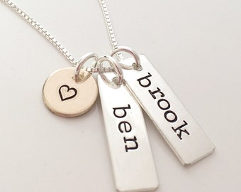 May include: A silver chain necklace featuring three dangling charms: a small circular disc stamped with a heart outline, and two rectangular silver bars stamped with the names ben and brook in lowercase black lettering.