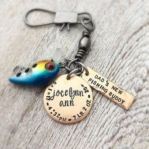 May include: A personalized fishing lure keychain with a blue, yellow, and black lure. The keychain includes a round tag with the name "Jocelynn Ann", birthdate, time, length, and weight. A rectangular tag reads "DAD'S NEW FISHING BUDDY".