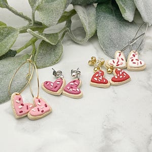 May include: Three pairs of heart-shaped cookie earrings with pink, red, and white frosting and sprinkles. The earrings are made of polymer clay and come in hoop and stud styles.
