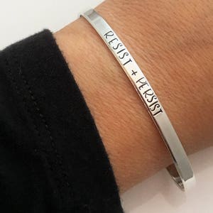 May include: A silver bangle bracelet with the words "Resist + Persist" engraved on it.