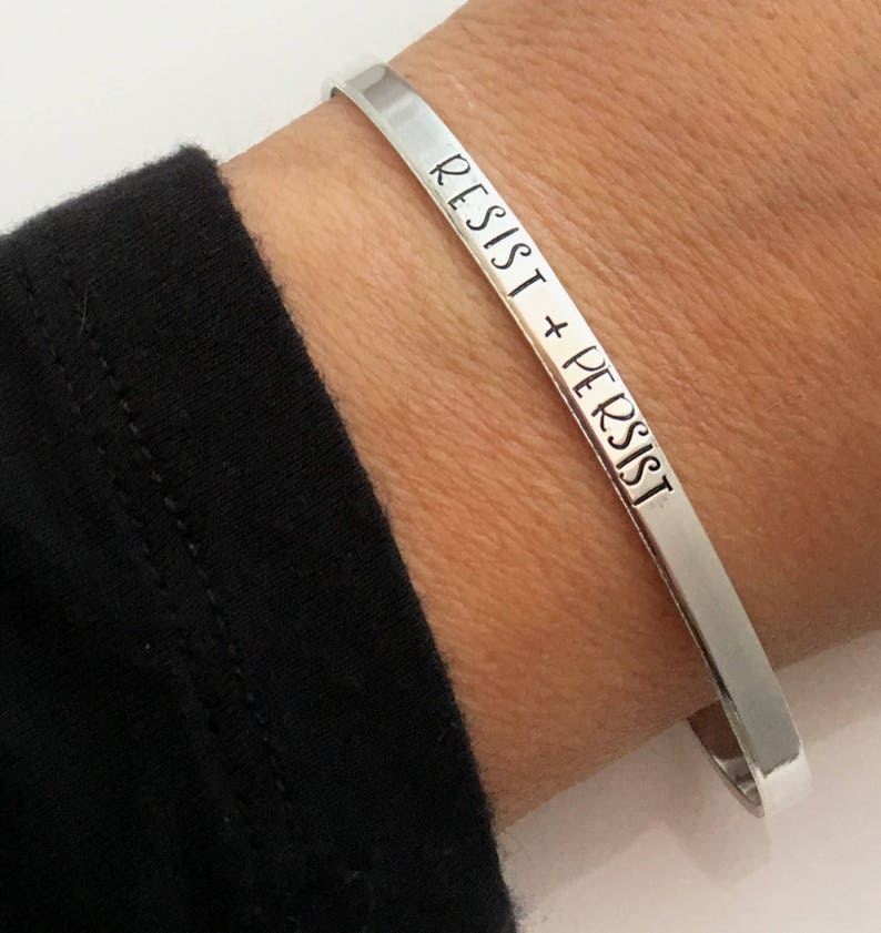 May include: A silver bangle bracelet with the words "Resist + Persist" engraved on it.