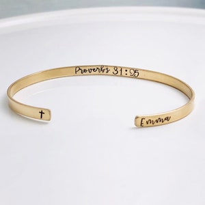 May include: A gold-colored cuff bracelet with the text "Proverbs 31:25" and the name "Emma" engraved. A small cross is on the opposite end. The bracelet is open-ended and sits on a white surface.