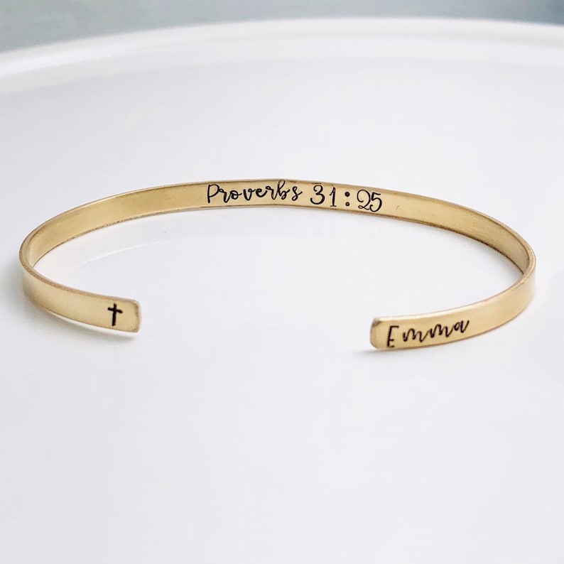 May include: A gold-colored cuff bracelet with the text "Proverbs 31:25" and the name "Emma" engraved. A small cross is on the opposite end. The bracelet is open-ended and sits on a white surface.