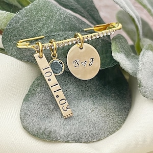 May include: A gold-colored safety pin with a row of clear crystals. It has three charms: a rectangular tag with the date "10.11.03", a small blue gemstone, and a round charm with the initials "B ♡ J". The pin is displayed on a green and white surface.