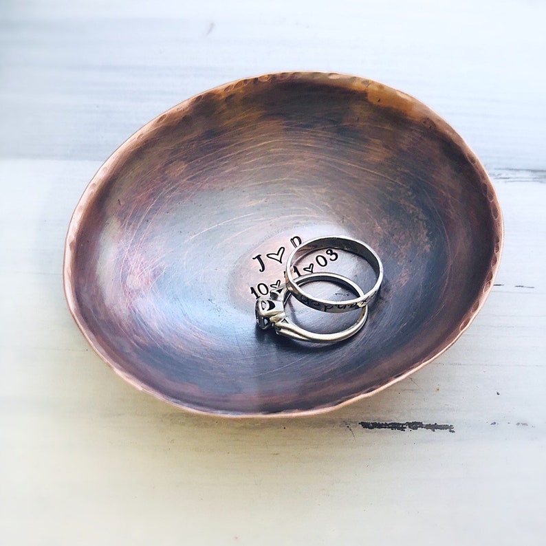 May include: A small, round, hammered copper bowl with a dark, burnished finish. Inside the bowl, the text J heart D and 10 heart 11 heart 03 is stamped. Two silver-toned metal rings are resting inside the bowl, one featuring a small stone setting.