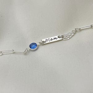 Op de afbeelding: Zilveren kettingarmband met een blauwe kristallen bedel en een zilveren bedel met de gravure "mom". De bedel is bevestigd aan een zilveren vleugelbedel.