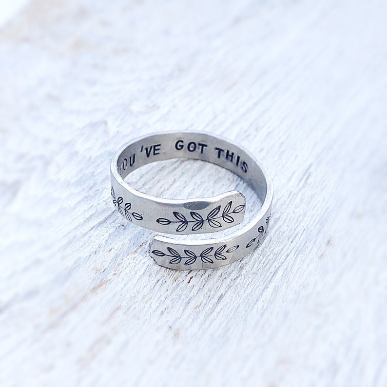 May include: A silver adjustable ring with the words "You've Got This" engraved on the band. The band is decorated with a repeating pattern of small leaves.