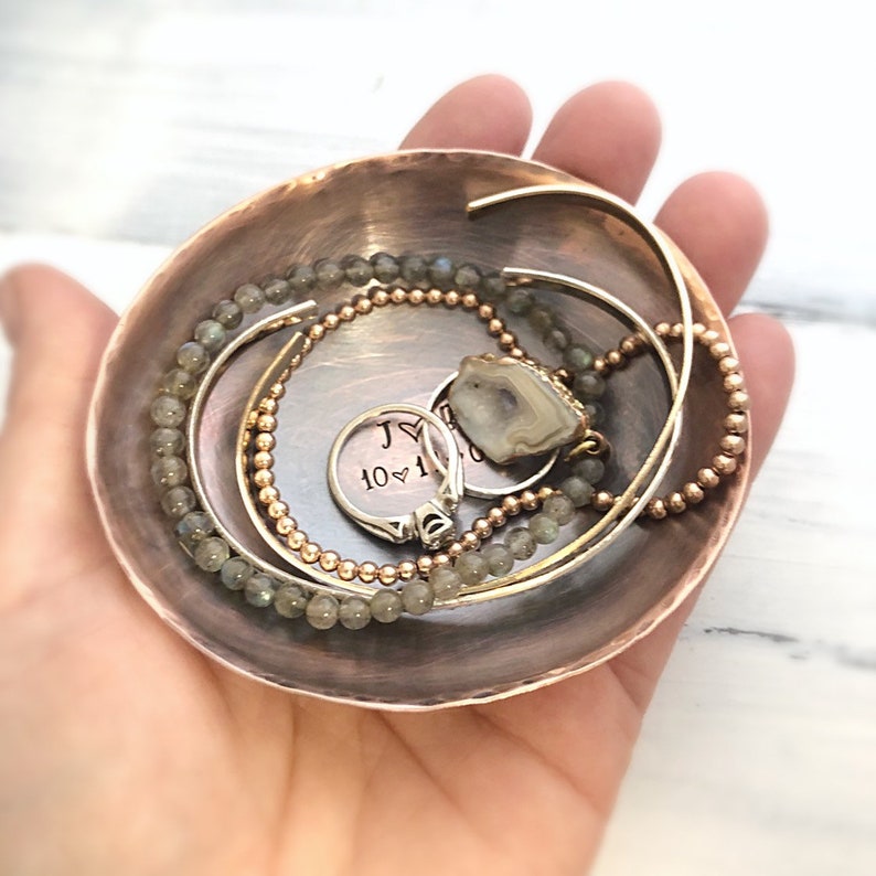 May include: A round, hammered copper trinket dish featuring the stamped text J heart 10 10 20. The dish contains a silver ring with a stone, a beaded bracelet with grey stones, a beaded bracelet with metallic beads, and a thin metal cuff.