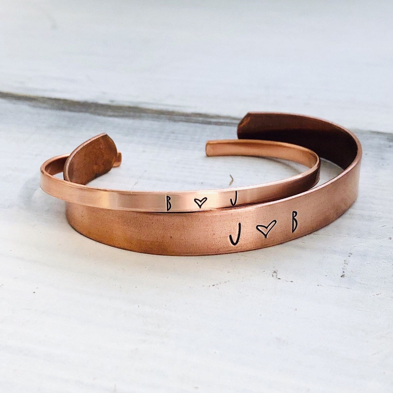 May include: Two copper bracelets with engraved initials and a heart symbol. The bracelets are stacked on top of each other. The top bracelet has the initials "B" and "J" with a heart symbol between them. The bottom bracelet has the initials "J" and "B" with a heart symbol between them.