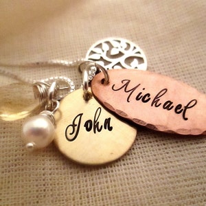 Puede incluir: Un collar personalizado con m&uacute;ltiples colgantes. El collar presenta una cadena plateada, una gema amarilla en forma de l&aacute;grima, una perla blanca y tres colgantes de metal. Dos colgantes est&aacute;n grabados con los nombres "John" y "Michael".
