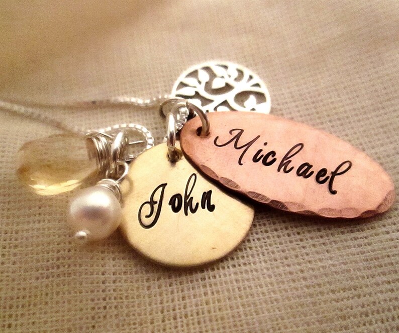 Puede incluir: Un collar personalizado con m&uacute;ltiples colgantes. El collar presenta una cadena plateada, una gema amarilla en forma de l&aacute;grima, una perla blanca y tres colgantes de metal. Dos colgantes est&aacute;n grabados con los nombres "John" y "Michael".