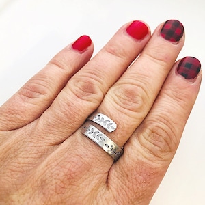 May include: A silver ring with a leaf design wrapped around the finger. The ring is worn on the middle finger of a hand with red nail polish.
