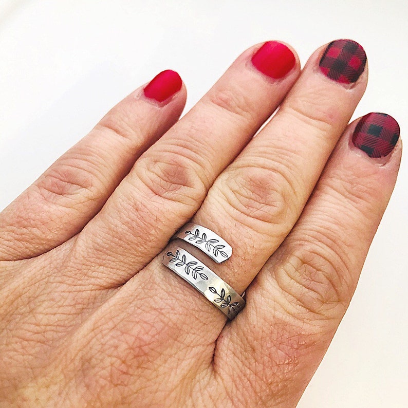 May include: A silver ring with a leaf design wrapped around the finger. The ring is worn on the middle finger of a hand with red nail polish.