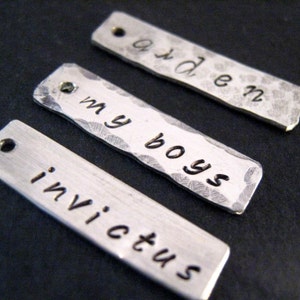 May include: Three rectangular, hammered metal tags with stamped text. The top tag reads "arden", the middle tag reads "my boys", and the bottom tag reads "invictus". Each tag has a small hole at one end.