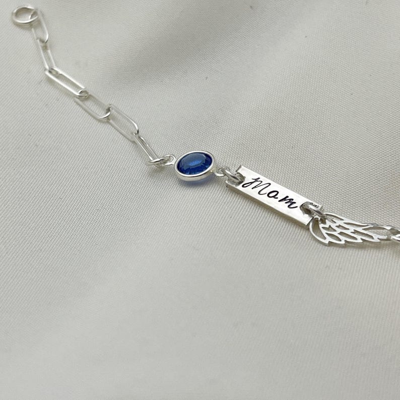 Op de afbeelding: Zilveren kettingarmband met een blauwe edelsteen, een bedel met de gravure "mam" en een vleugelbedel.