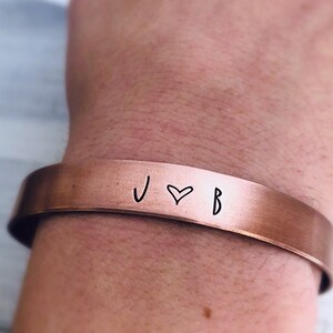 May include: A copper cuff bracelet with the initials "J" and "B" separated by a heart symbol.