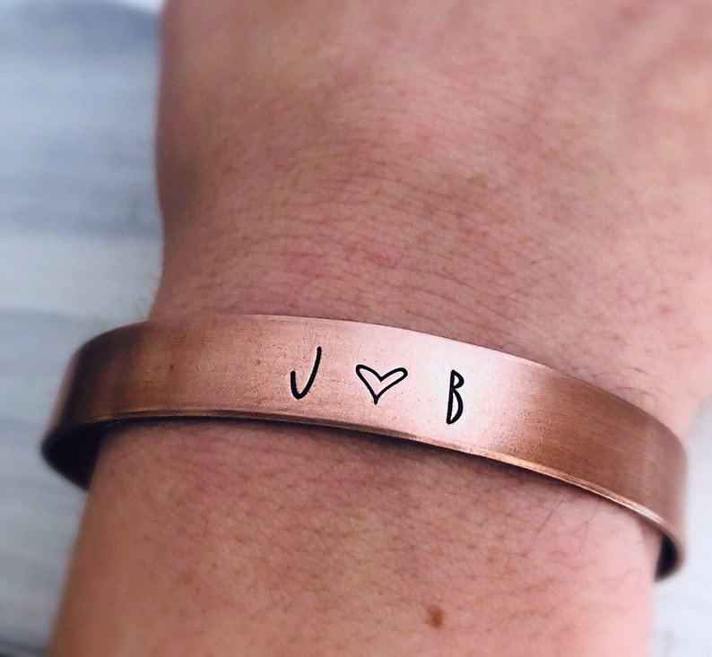 May include: A copper cuff bracelet with the initials "J" and "B" separated by a heart symbol.