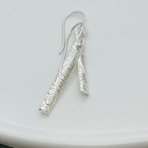 May include: Silver dangle earrings with a floral design. The earrings are made of two pieces of silver that are connected by a loop. The top piece is longer and has a hook for attaching to the ear. The bottom piece is shorter and has a floral design.