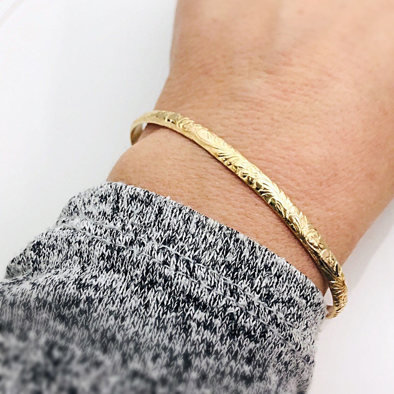 May include: A gold bangle bracelet with an intricate floral design. The bracelet is thin and delicate, and it is shown on a person's wrist.