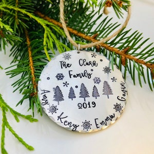 May include: A silver Christmas ornament with the text "The Clark Family 2019" and the names "Amy, Kenny, Emma, Gavin" around the edge. The ornament features five stylized pine trees and snowflakes.