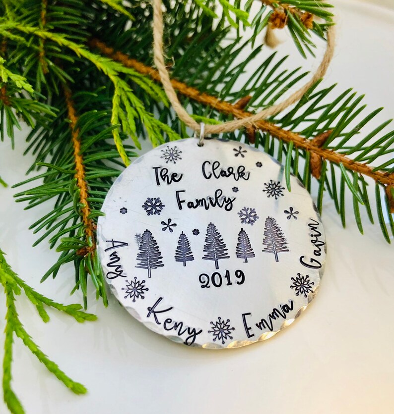 May include: A silver Christmas ornament with the text "The Clark Family 2019" and the names "Amy, Kenny, Emma, Gavin" around the edge. The ornament features five stylized pine trees and snowflakes.