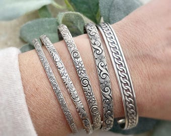 May include: Five silver bracelets with intricate floral and chain designs. The bracelets are stacked on a person's wrist.