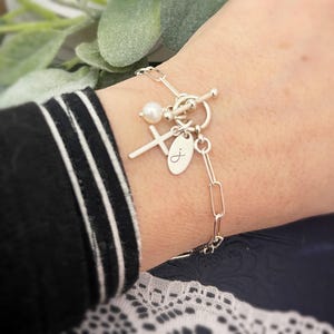 May include: A silver charm bracelet with a cross, a pearl, and a personalized oval charm with the letter "J". The bracelet has a toggle clasp.
