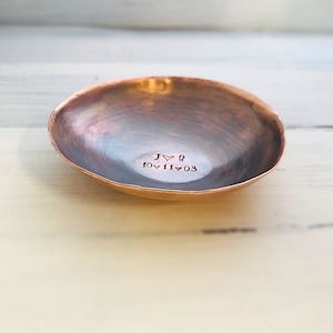 May include: A small, round, hammered copper bowl with a dark, burnished finish. The interior is stamped with the text J heart B and the date 10 heart 11 heart 03. The bowl has a shallow, concave shape with a slightly irregular, organic rim.