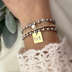 May include: A stack of beaded bracelets in silver and gold tones. One bracelet features a small gold tag with the letters "R" and "K" and a heart symbol. Another bracelet has a small gold heart charm. A pearl bead is also visible.
