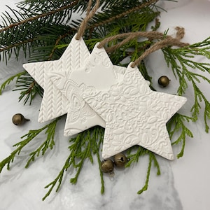 May include: Three white ceramic star ornaments with embossed designs. The stars are hanging from twine and are surrounded by green pine branches.