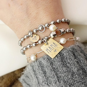 May include: A silver chain bracelet with a gold charm that says "MAMA" with a small heart. The bracelet also has a small heart charm and two white pearl charms.