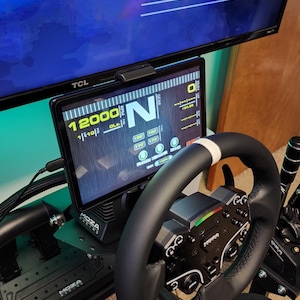 May include: A black racing wheel with a white stripe, a black and green shifter, and a black and silver racing pedal set. A black tablet with a yellow screen displays the text "12000 RPM" and "N". The tablet is mounted on a black stand. The text "Progresso" and "ANO 23.3% MES: 83.9% DIA: 73.7%" is visible on a blue screen in the background.