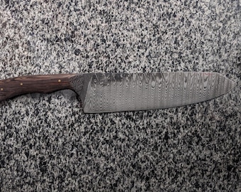 May include: A long, sharp kitchen knife with a dark wood handle and a wavy, patterned blade. The knife is lying on a gray, speckled surface.