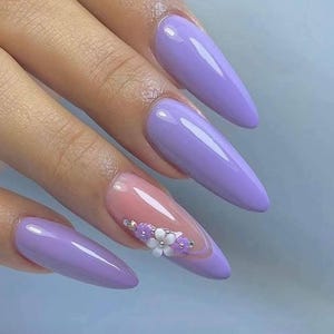 Puede incluir: Primer plano de uñas largas y puntiagudas pintadas de un tono lila claro. Una uña presenta un delicado diseño de flores blancas y moradas.