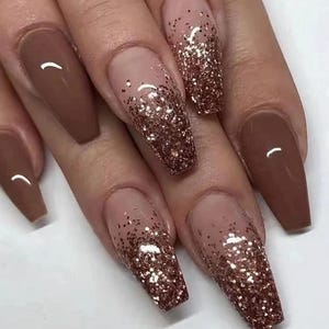 Puede incluir: Uñas largas en forma de almendra con una base marrón y un degradado de purpurina dorada en las puntas.