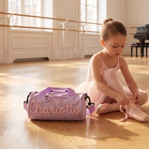 May include: A light purple cylindrical duffel bag featuring the name Charlotte embroidered in a matching script font on the front, equipped with a zipper closure, two top handles, and a detachable shoulder strap with black hardware.
