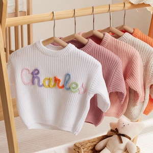 May include: A wooden clothing rack displays several pastel-colored sweaters and a straw hat with a pink bow. A white sweater with the name "Charles" embroidered in rainbow colors hangs prominently. A shelf holds baby shoes and a stuffed bear.