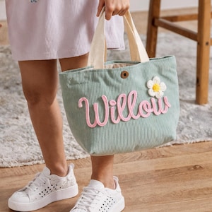 Può includere: Una borsa tote in velluto a coste verde chiaro con il nome "Willow" in corsivo rosa. La borsa ha un manico in tessuto bianco e una piccola applicazione floreale bianca e gialla. La persona indossa scarpe da ginnastica bianche.