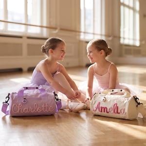 May include: Two cylindrical duffel bags with adjustable shoulder straps and top handles. One bag is light purple with the name Charlotte embroidered in pink, and the other is off-white with the name Amelia embroidered in pink and purple.