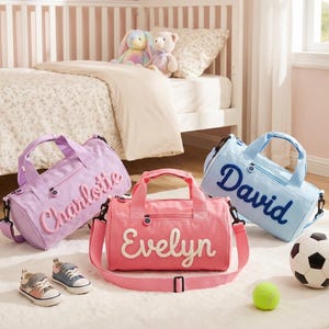 May include: Three small duffel bags in light purple, pink, and light blue. Each bag features a top zipper, two handles, and an adjustable shoulder strap. The names Charlotte, Evelyn, and David are embroidered in large, raised script letters on the front.