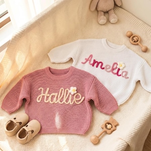 Op de afbeelding: Twee baby truien, één roze en één wit, met geborduurde namen en bloemaccenten. De roze trui zegt "Hallie" en de witte trui zegt "Amelia". Een paar beige babyschoenen en houten speelgoed zijn ook aanwezig.