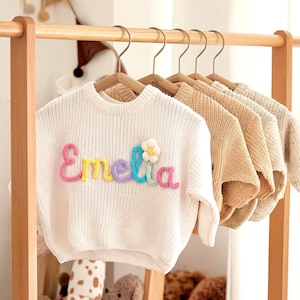Op de afbeelding: Een witte gebreide trui met de naam "Emelia" in kleurrijke letters en een kleine bloemapplicatie. Verschillende andere truien in diverse neutrale kleuren hangen aan een houten rek.