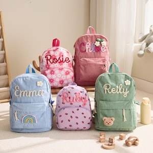 May include: Five personalized backpacks in various colors, including pink, blue, and green. Each backpack features a name embroidered on the front, along with decorative elements like flowers, rainbows, and a bear. A wooden bookshelf and toys are in the background.