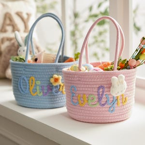 以下が含まれることがあります： 2つのイースターバスケットが並んでいます。1つは水色、もう1つはピンク色です。水色のバスケットにはカラフルな糸で「Oliver」という名前が刺繍されており、ピンク色のバスケットには「Evelyn」と刺繍されています。どちらもイースターのお菓子や装飾品でいっぱいです。