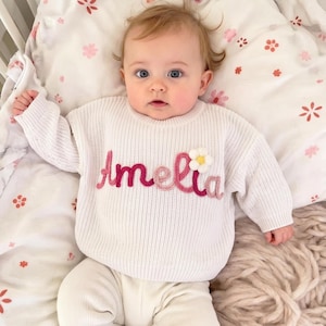 May include: A baby in a white knitted sweater embroidered with the name "Amelia" in pink and red, and a small flower. The baby wears white trousers and lies on a floral patterned blanket.