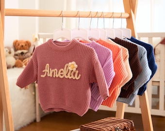 May include: A pink knitted sweater with a white flower and the name "Amelia" embroidered on the front. The sweater is hanging on a wooden rack with other sweaters in various colors.